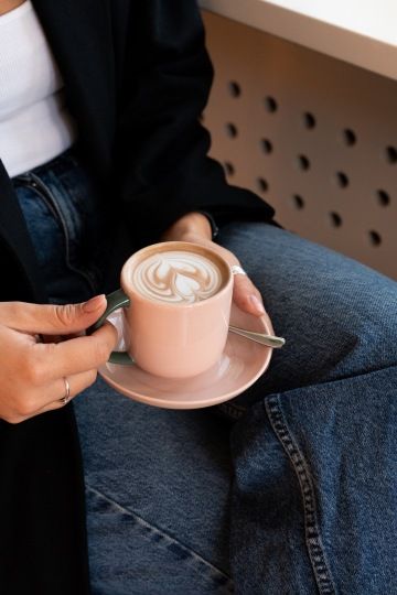 Osoba drží šálek cappuccina s latte art, oblečená v džínách a černém saku.