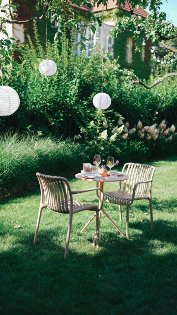 Zahradní stůl se dvěma židlemi, prostřený na letní večeři, lemovaný lampióny a zelenými rostlinami na svěží trávě.