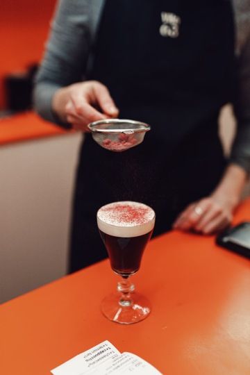 Osoba sype červený prášek přes sítko na černou kávu v průhledné sklenici.